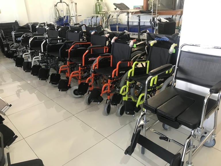 Multiple wheelchairs lined up in rows, including black, orange, and yellow models in an indoor facility.